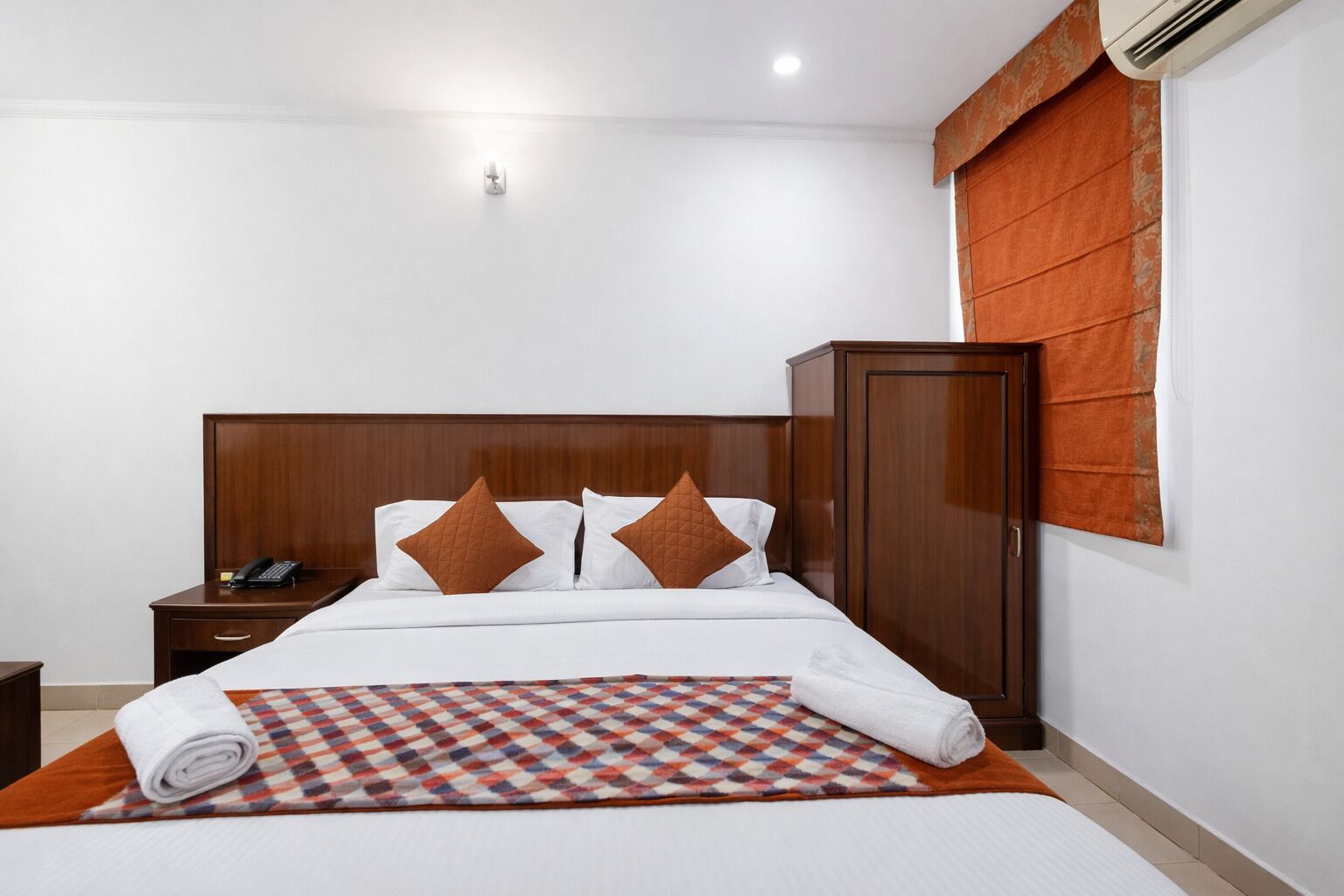 Quarto de hotel com cabeceira de madeira, cama com lençóis brancos e almofadas castanhas, telefone em uma mesa de cabeceira, tapete estampado e unidade de ar condicionado.