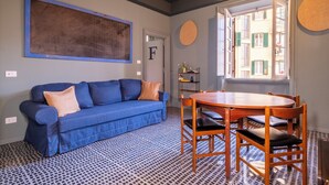 A room with a blue sofa, a wooden dining table with chairs, a patterned floor, a large abstract painting, and a view through an open window. - HT Vatican cozy rooms & apartments (Rome)