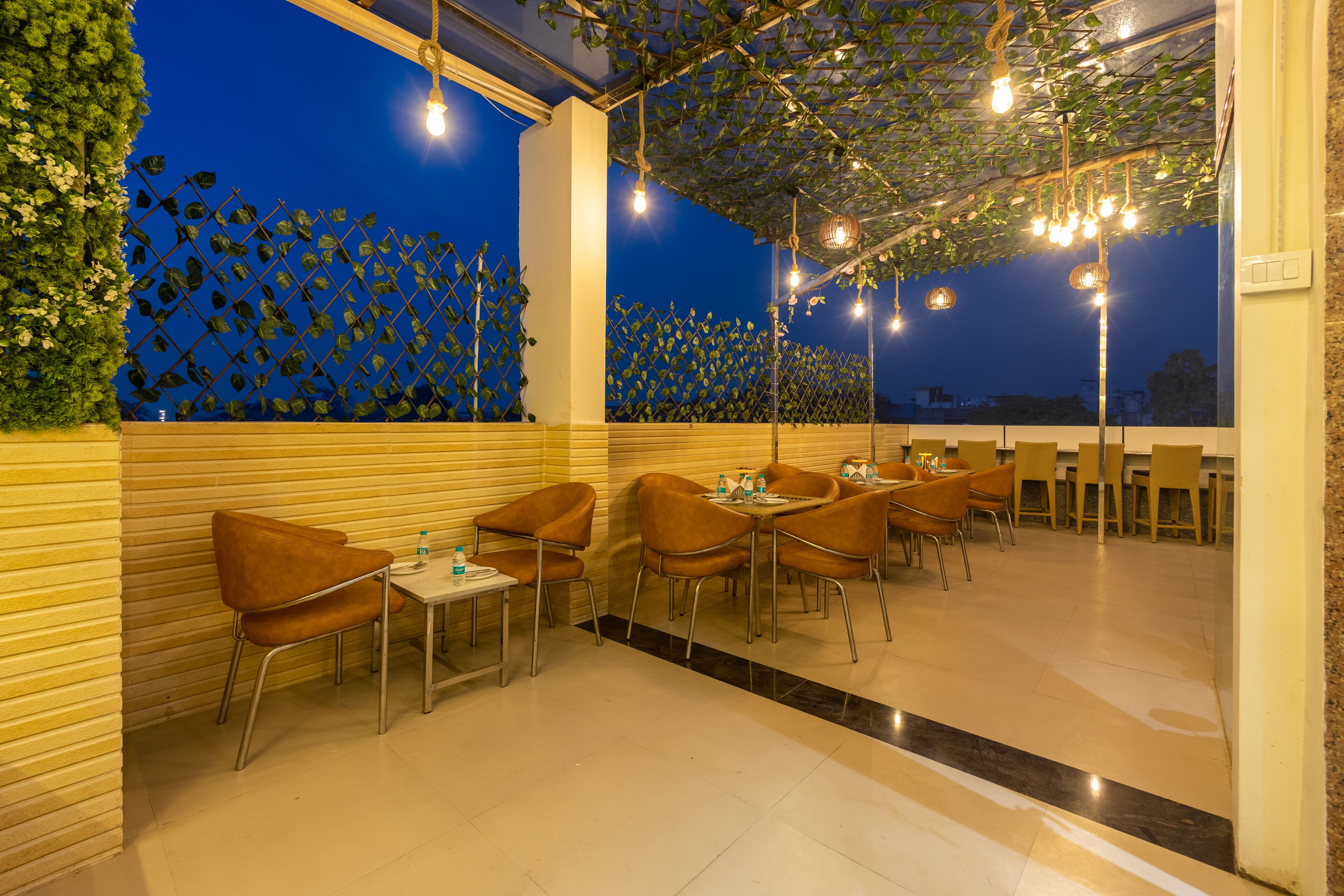 An outdoor dining area with tables and chairs, a leafy wall, and string lights.