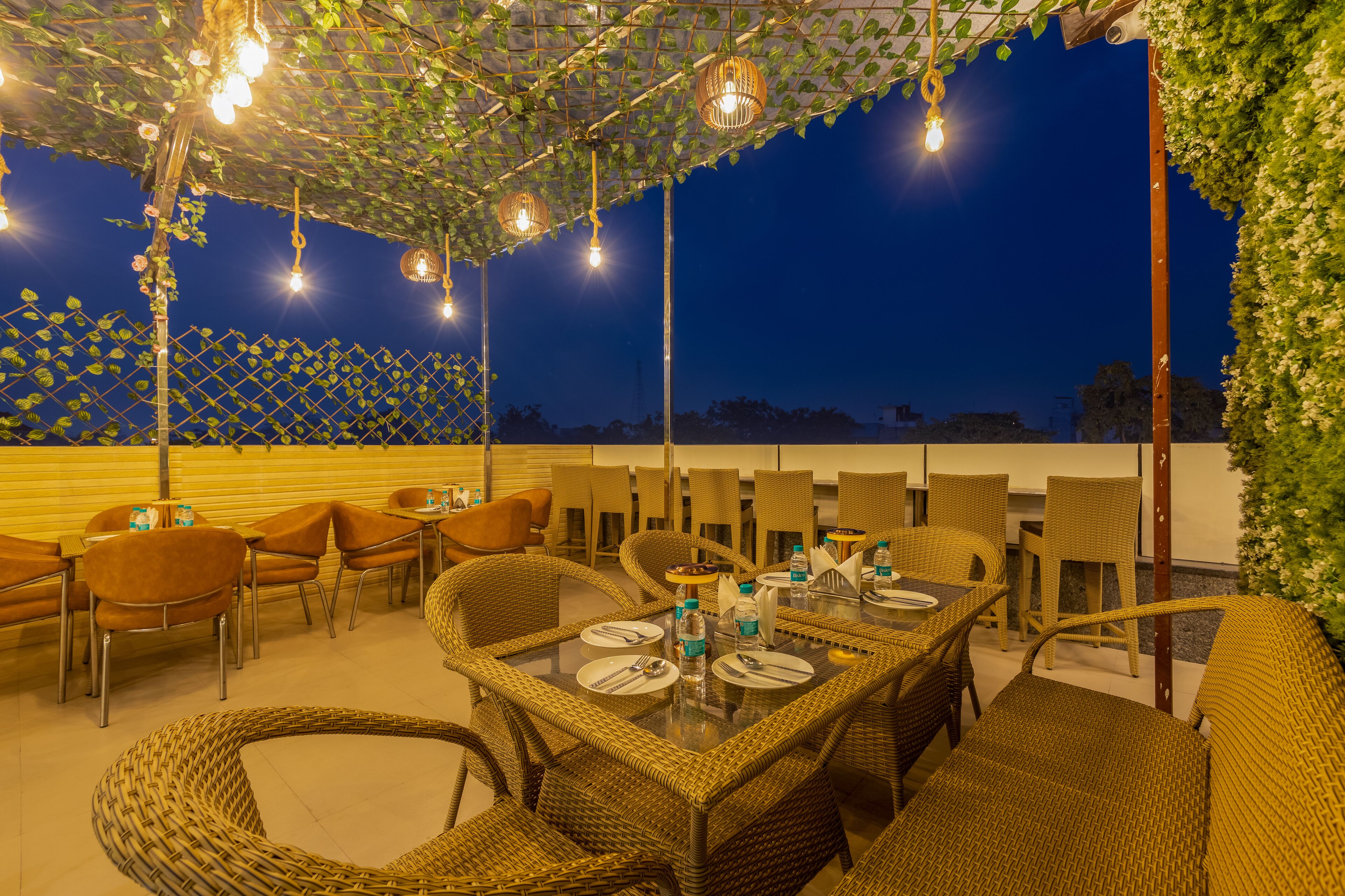 Un área de comedor al aire libre con muebles de mimbre e iluminación colgante, frente a un cielo nocturno.