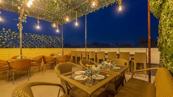 Un área de comedor al aire libre con muebles de mimbre e iluminación colgante, frente a un cielo nocturno.