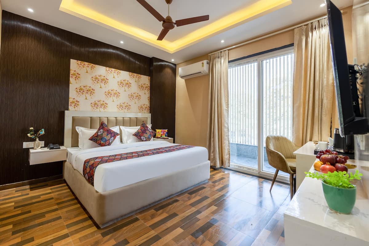A modern hotel room with a large bed, a ceiling fan, a flat-screen TV, a dining area with a fruit bowl, and a large window with curtains.