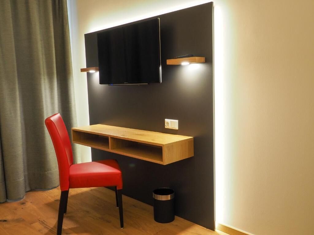 Quarto moderno com escrivaninha de madeira, cadeira vermelha, lixeira preta e televisão na parede.