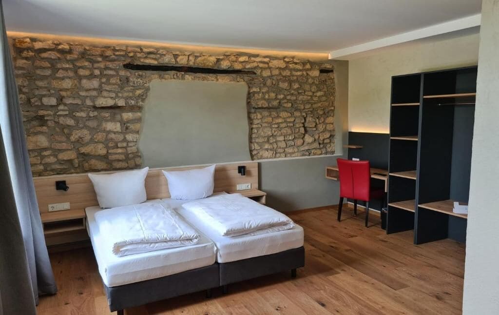 Un dormitorio con pared de piedra, cama con ropa de cama blanca, una silla roja y un estante negro.