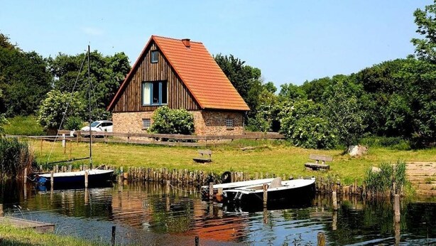 A rustic house with a sloped roof and wooden exterior, situated by a calm water body with boats docked. - Condé RE-10, Germany (Stolpe auf Usedom)