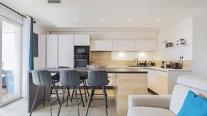 A modern kitchen with a central island, bar stools, and a sofa with a blue pillow. - Q&R Renting- Apartments and Suites- Parking AC Mer (Nice)
