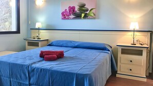 Une chambre avec une couvre-lit bleue, deux tables de chevet avec des lampes, un tableau représentant des fleurs et des pierres accroché au mur, et un mur en pierres apparentes.