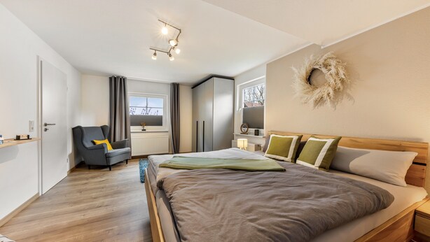 Ein modernes Schlafzimmer mit einem großen Bett, einem grauen Sessel, einem Kleiderschrank und einem Fenster mit Vorhängen.