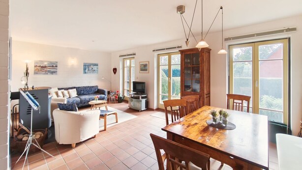 Un salón amplio con sofá, mesa de comedor y televisor. Hay ventanas grandes y un mueble bar con puertas de cristal.