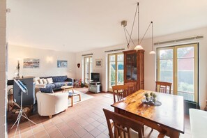 A spacious living room with a sofa, a dining table, and a TV. There are large windows and a cabinet with glass doors. - Apartment Merkel - Comfortable apartment in Koserow with terrace, (Koserow)