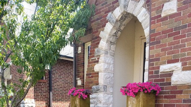 A brick building with a stone archway entrance, steps leading up to a door, and large flower pots with pink flowers on the steps. - Charming 3BR STL Home • Sleeps 6 • Free Parking (St. Louis)