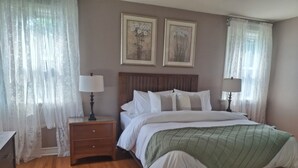 A bedroom with a wooden floor, a bed with white bedding, two bedside lamps, a nightstand, and a wooden bench. - Charming 3BR STL Home • Sleeps 6 • Free Parking (St. Louis)