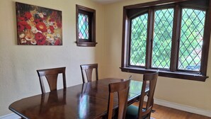 A dining room with a wooden table and chairs, a painting on the wall, and a window with a view of greenery. - Charming 3BR STL Home • Sleeps 6 • Free Parking (St. Louis)