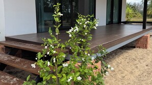 Ein moderner Außenbereich mit einer Holzterrasse, einer kleinen Pflanze mit weißen Blüten und einem Gebäude mit großen Fenstern und einer überdachten Terrasse.