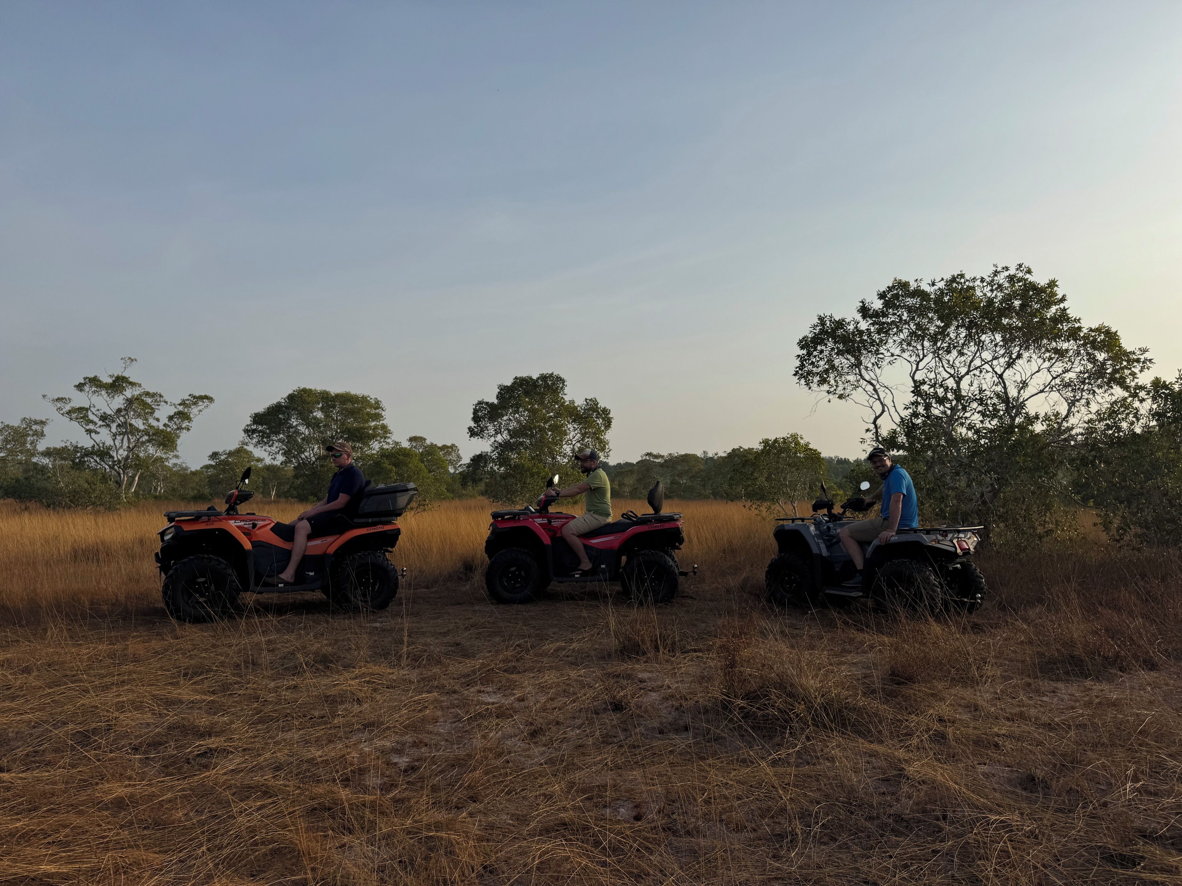 Three people riding ATVs in a dry grass field with trees in the background.