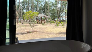Ein Zimmer mit einer großen Badewanne, einer Holzbalkendecke und Blick auf eine tropische Außenanlage mit Bäumen und einem Reetdachgebäude.