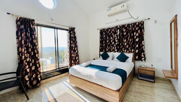 A bedroom with a large bed, a nightstand, a chair, and a window with curtains. - JK Valley by Shree Stays (Mahabaleshwar)