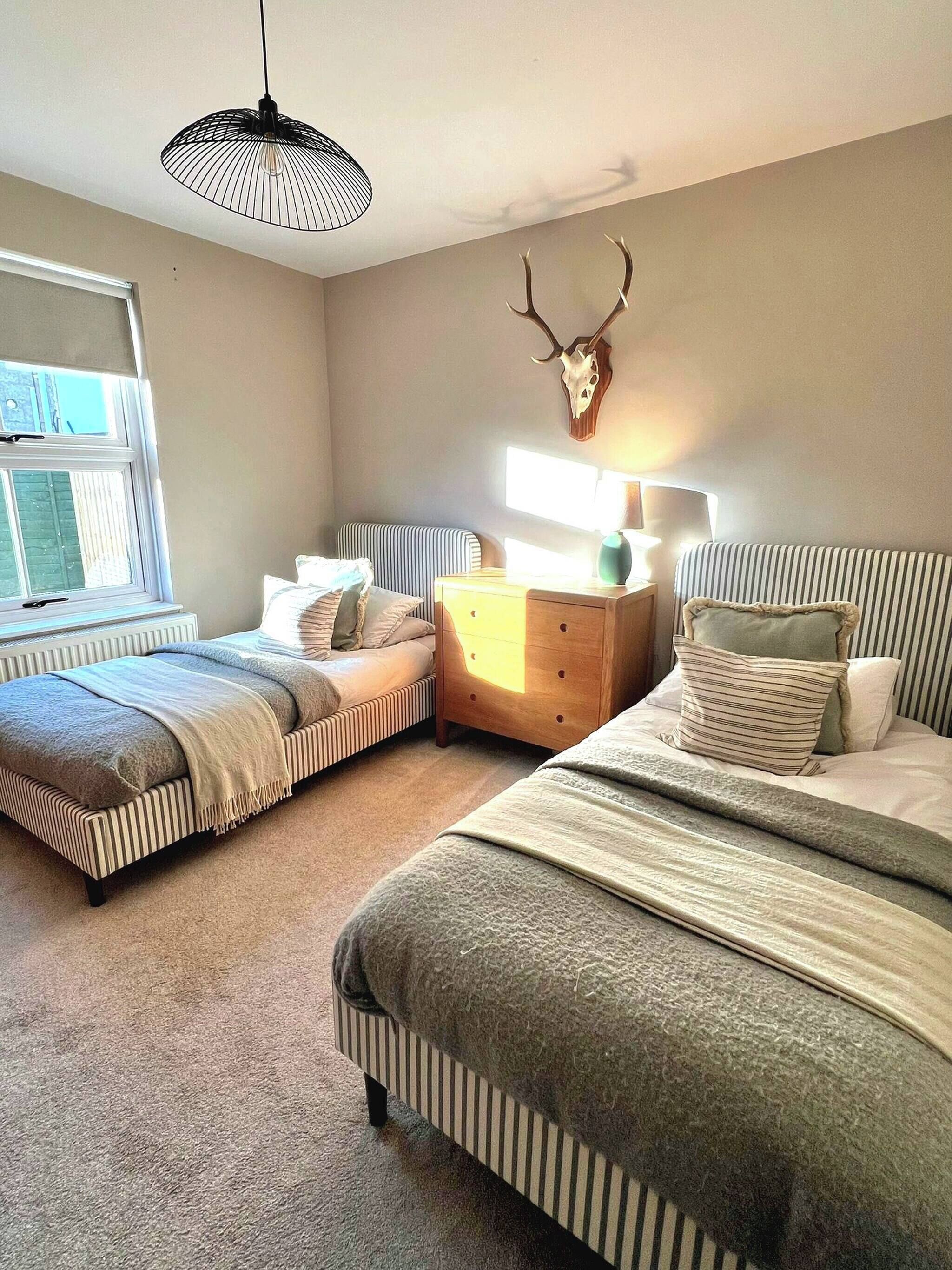 Une chambre avec deux lits, une table de chevet en bois, une lampe, un crâne de cerf accroché au mur et une fenêtre avec des stores.
