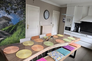 A dining area with a long table, chairs, a clock, and a kitchen with appliances.