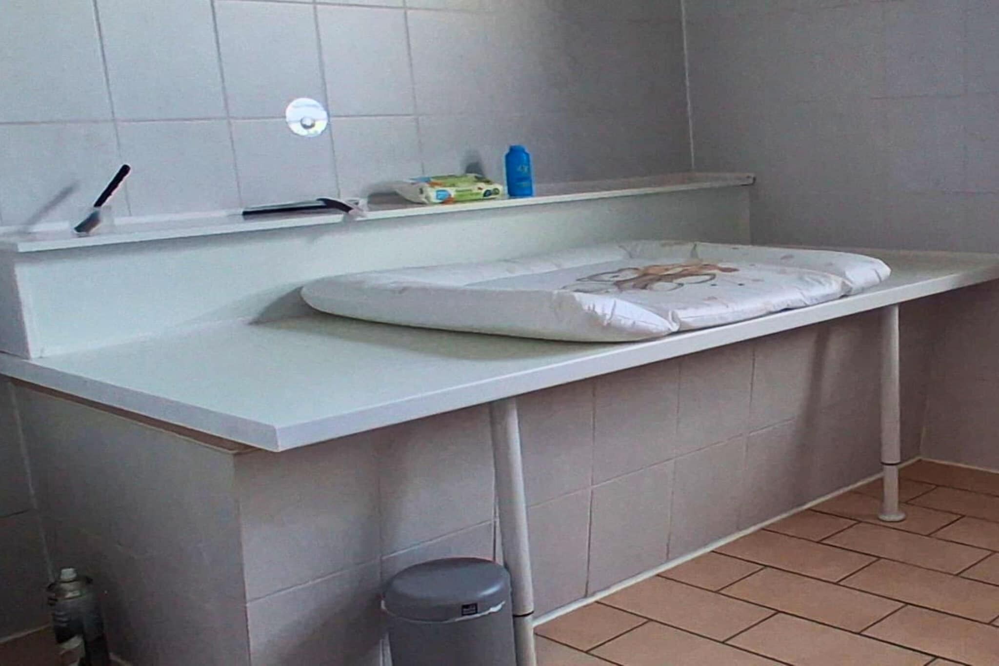 Ein Badezimmer mit weißer Ablage, Waschbecken und Toilette. Neben der Toilette steht ein Mülleimer und über dem Waschbecken befindet sich ein Ablagefach mit einer Zahnbürste und einer Flasche.
