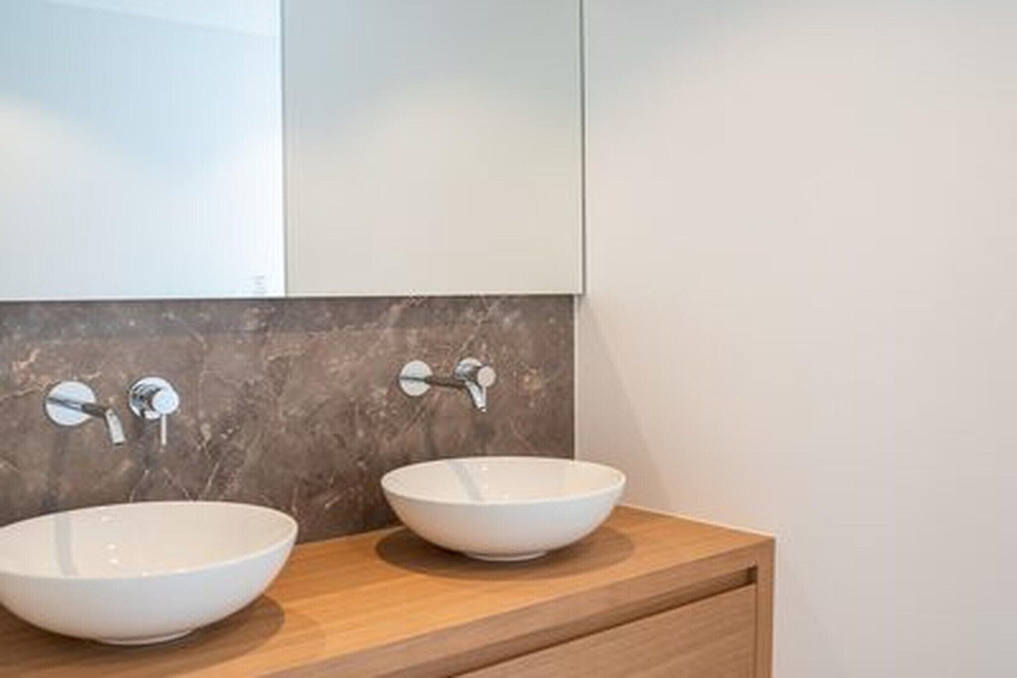 Une salle de bain avec deux vasques blanches posées sur un meuble-lavabo en bois, un grand miroir au-dessus et un pare-baigneoire en marbre.