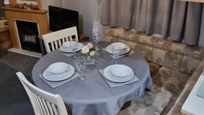 Een eettafel gedekt voor vier personen, met witte borden en glazen, een vaas met bloemen, en een open haard op de achtergrond.
