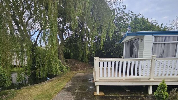 Een stacaravan met een witte buitenkant en een balkon, omringd door bomen en een klein gazon.