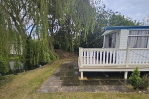 A mobile home with a white exterior and a balcony, surrounded by trees and a small grassy area. - Caravan - Sleeps 6 - Pets Ok - Parking (Skegness)