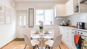 A modern kitchen with white cabinets, a round dining table with four chairs, and a window with curtains. - New! Delightful 2BR Flat in Woolwich (Greater London)