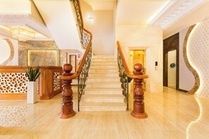 A grand staircase with marble steps and wooden balusters, leading to a landing with a reception desk and a potted plant.