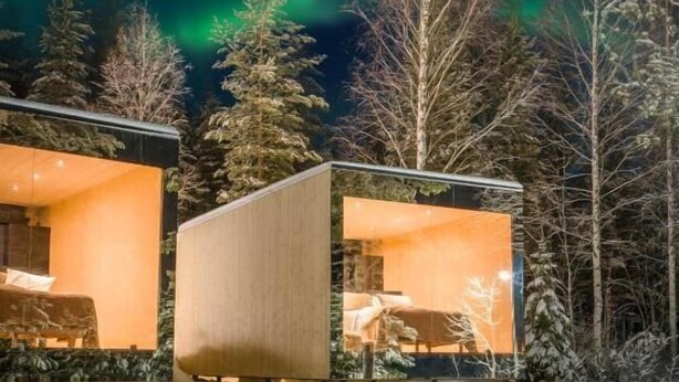 Una cabaña moderna con fachada de vidrio, rodeada de suelo y árboles cubiertos de nieve, bajo un cielo nocturno con auroras boreales.