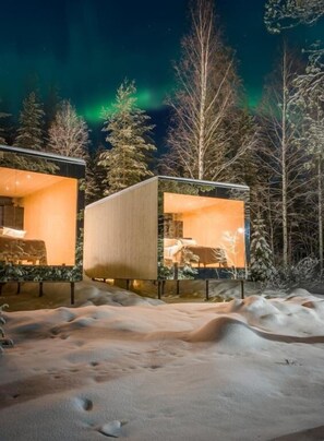 Eine moderne Holzhütte mit Glasfassade, umgeben von verschneitem Boden und Bäumen, unter einem Nachthimmel mit Polarlichtern.