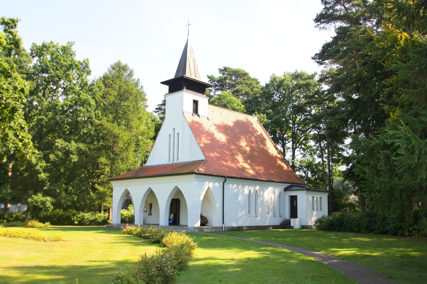 A church with a steeple, arched windows, and a red roof, surrounded by greenery.