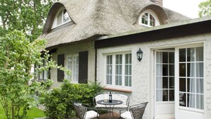 Ein Haus mit einem Reetdach und weißer Fassade, eine Terrasse mit rundem Tisch und Stühlen sowie eine Tür mit Glasfeldern.