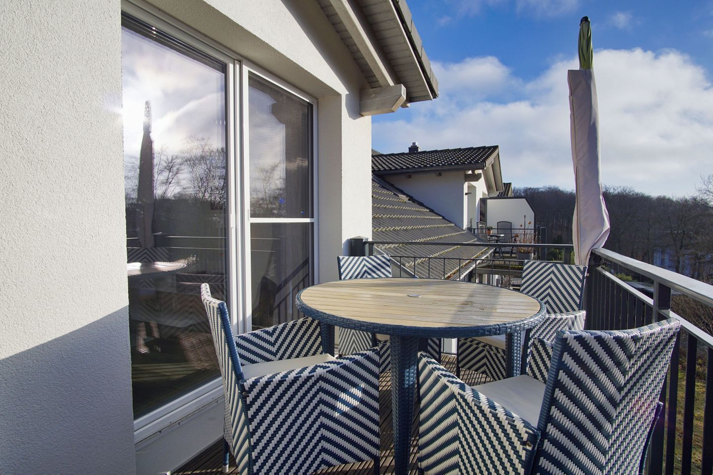 Een balkon met een ronde tafel en stoelen, grote ramen en uitzicht op een gebouw en bomen.