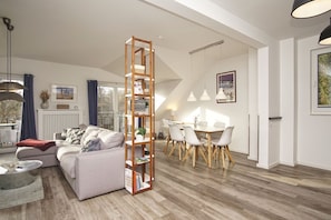 Ein modernes Wohnzimmer mit einer Couch, einem hohen Holzregal und einem Essbereich mit Tisch und Stühlen.