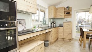 A modern kitchen with wooden cabinets, a built-in oven, a sink, and a dining area with a table and chairs. - Rügen vacation near the beach - Landhaus Wilkens Bungalow Nordlicht - Country house (Baabe)
