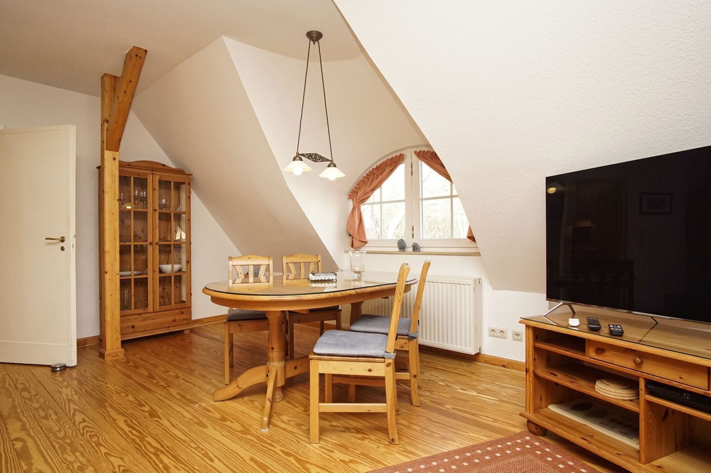 A cozy attic room with a wooden dining table, chairs, a cabinet with glass doors, a television, and a window with curtains.