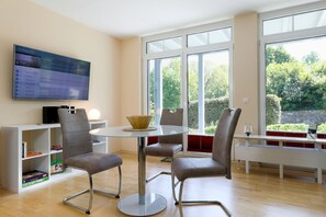 Ein modernes Wohnzimmer mit Glastisch, vier grauen Stühlen, einem Fernseher an der Wand und Blick ins Grüne.