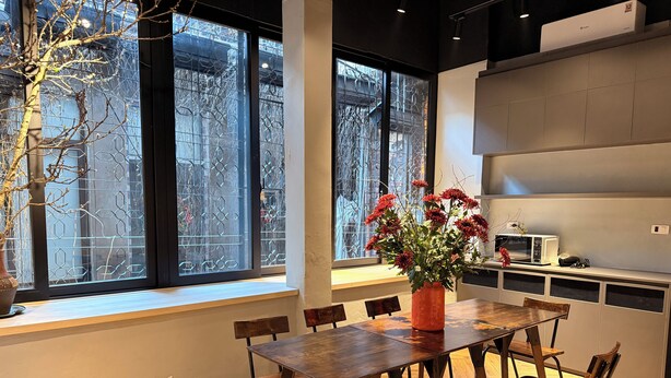 Et moderne spiseområde med et træbord og stole, en vase med røde blomster og store vinduer med udsigt til træer og bygninger.