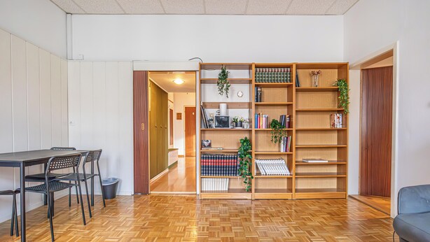 A modern living room with a wooden floor, a dining area with chairs, a bookshelf, and a grey sofa.