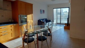 A modern kitchen with wooden cabinets, a glass dining table, and chairs. There is a sofa and a large window with blinds in the living area.