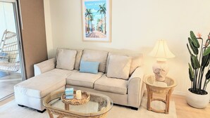 A living room with a beige sectional sofa, a glass coffee table, a wicker side table with a lamp, and a large potted plant. - Retro coastal Fort Myers retreat with private yard, spa & fire pit. Two bedrooms, two baths, sun room with fitness wall, screened patio, and easy access to RSW, FGCU, shopping, and beaches. (Fort Myers)