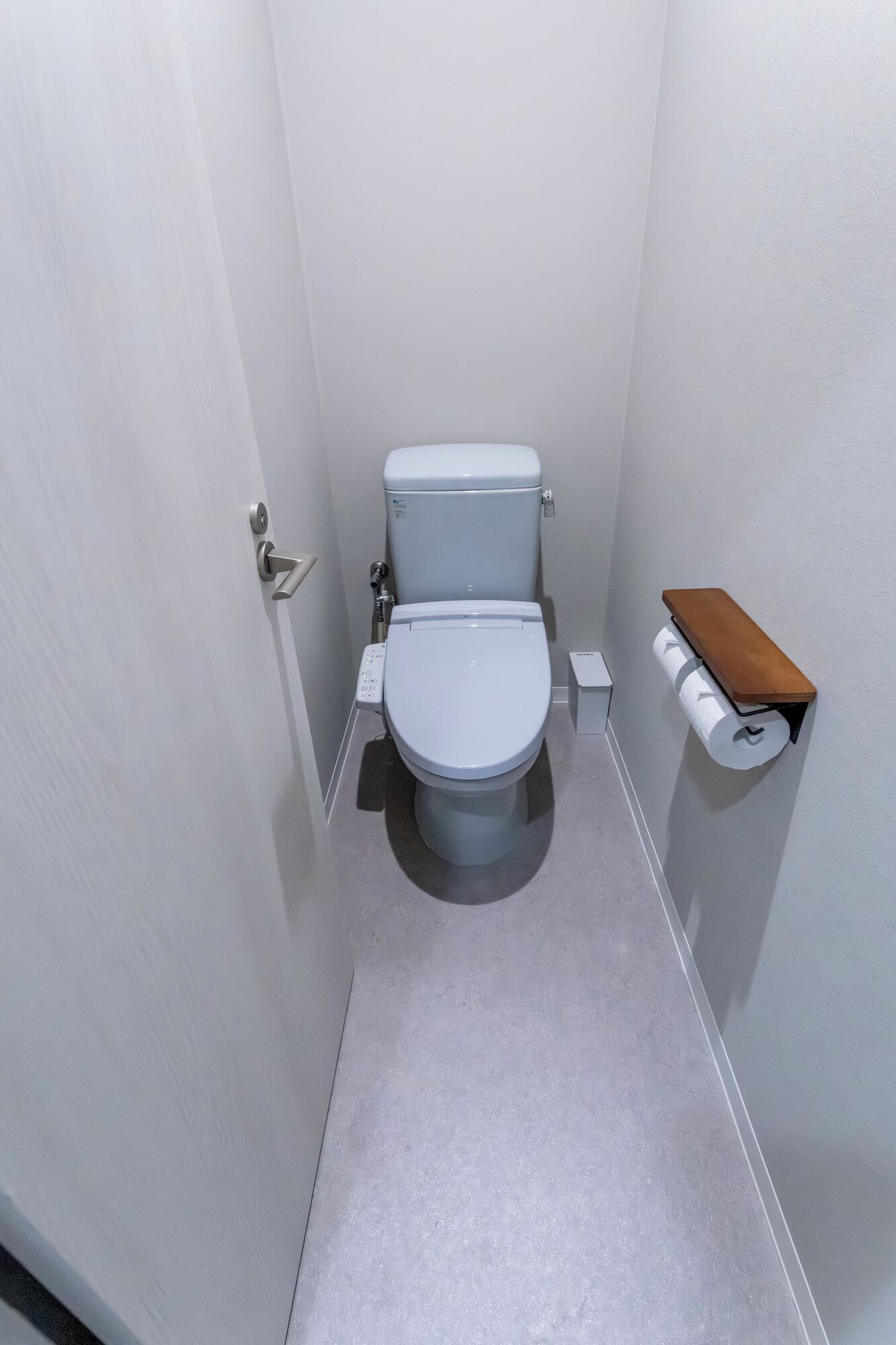 Un baño pequeño y estrecho con inodoro blanco, dispensador de papel higiénico y una estantería pequeña.