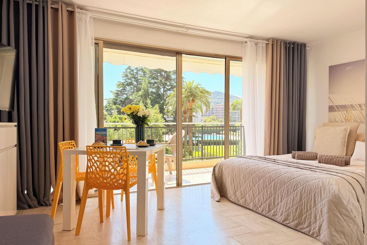 A hotel room with a balcony, a bed, a dining table with chairs, and a view of the outdoors.