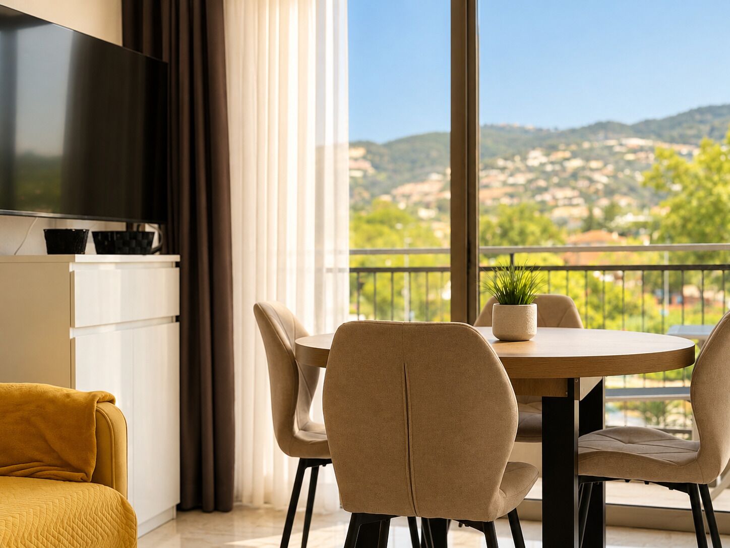 A modern living room with a TV, a sofa, a dining table with chairs, and a balcony with a view of the countryside.