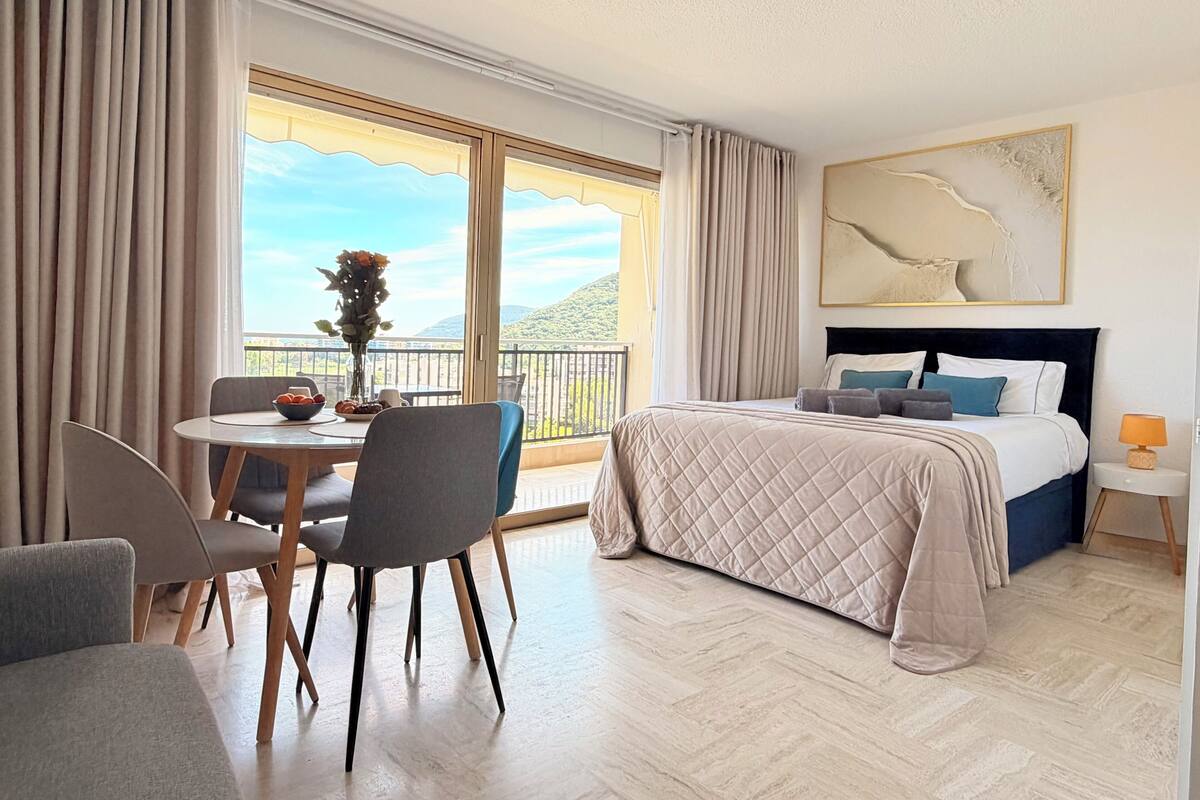 A hotel room with a bed, a dining table with chairs, a vase of flowers, and a balcony with curtains.