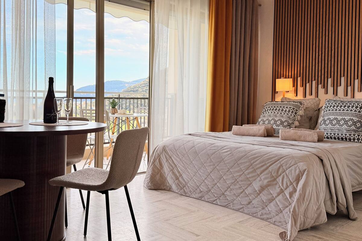 A hotel room with a bed, a table with a bottle and glasses, a chair, and a balcony with a view.
