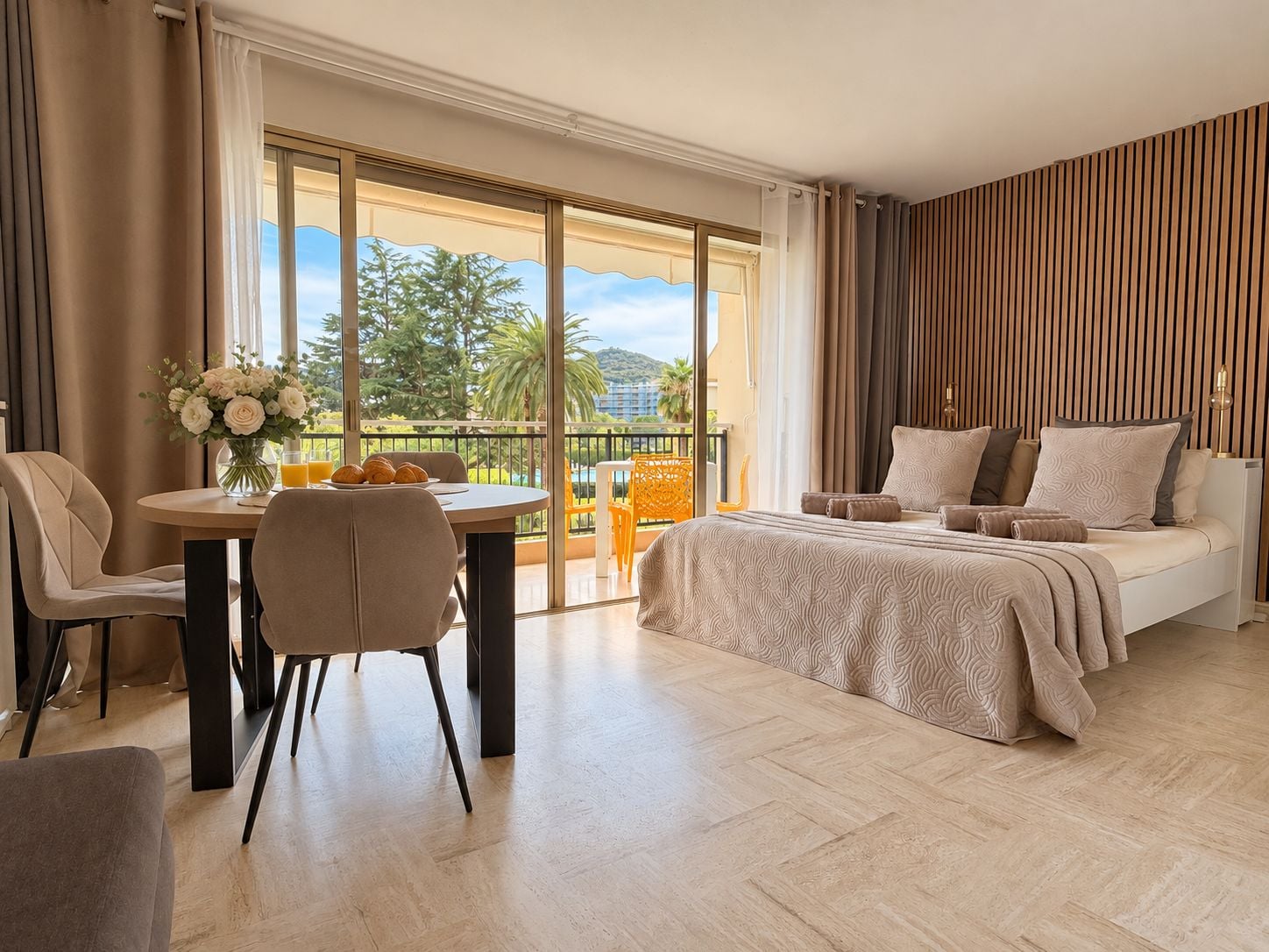 A hotel room with a bed, a dining table with chairs, a balcony with a view of palm trees and a building, and a large window.