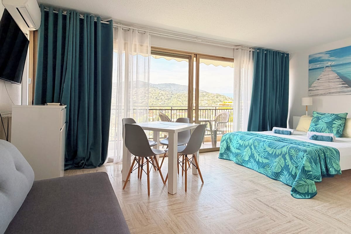A hotel room with a balcony, a dining table with chairs, a bed with a patterned bedspread, and a TV.